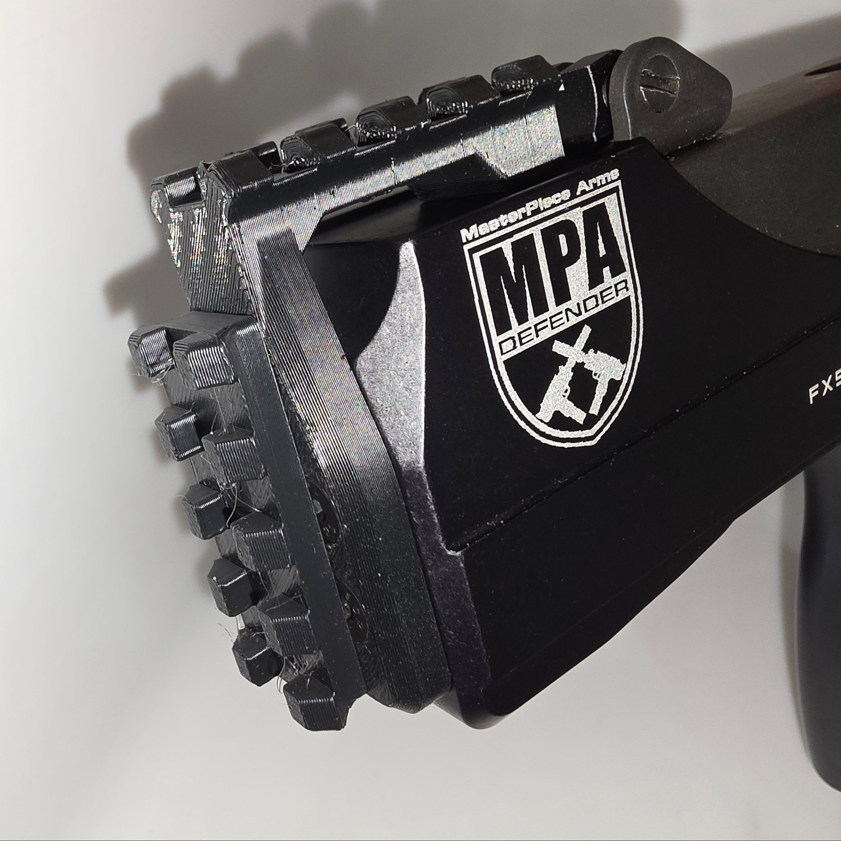 Close-up of a black MasterPiece Arms MPA Defender FX59042 paintball barrel tip with a white logo decal, held by a hand against a plain white background.