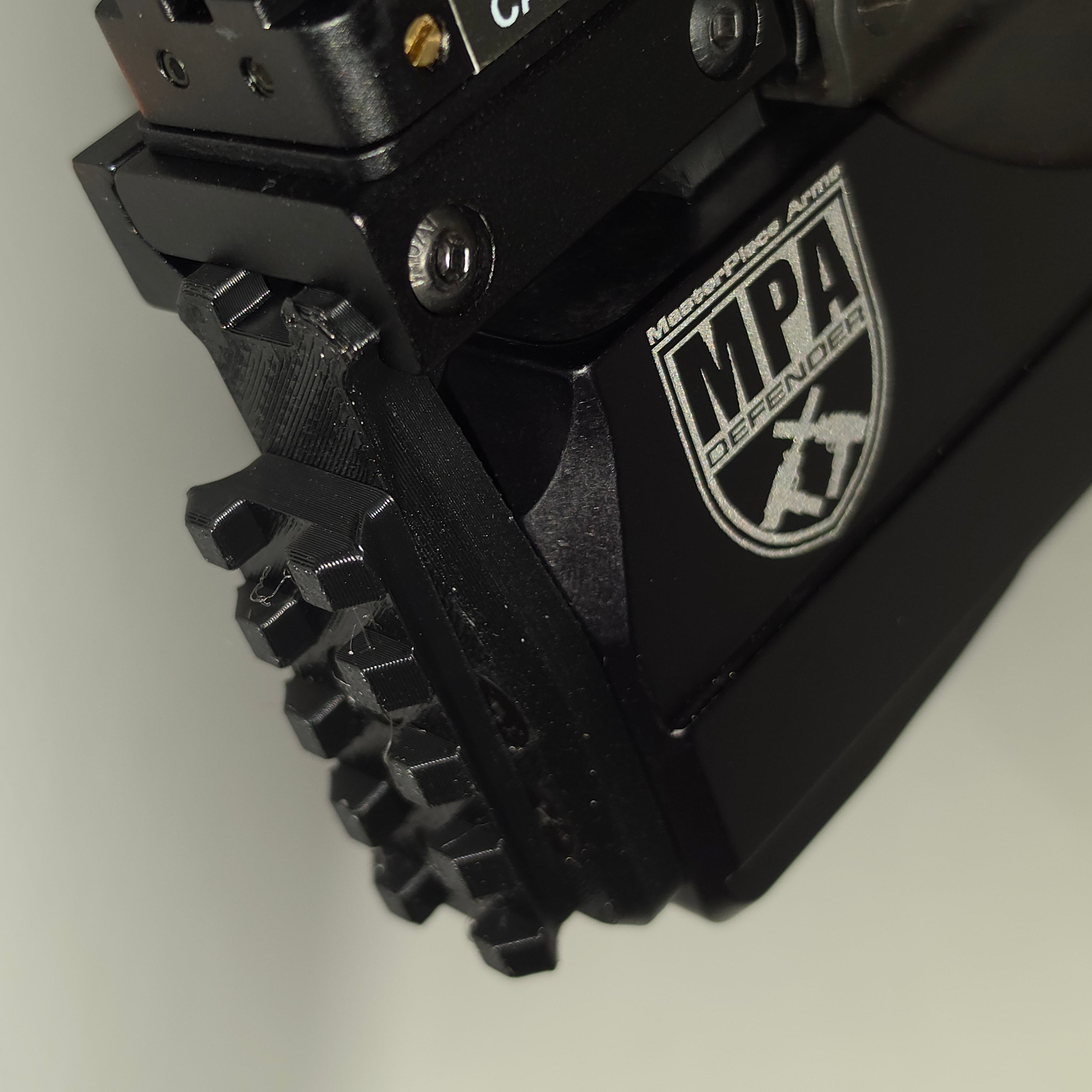 Close-up of a black tactical firearm accessory with a textured grip and a visible MPA Defender logo on a light gray background.