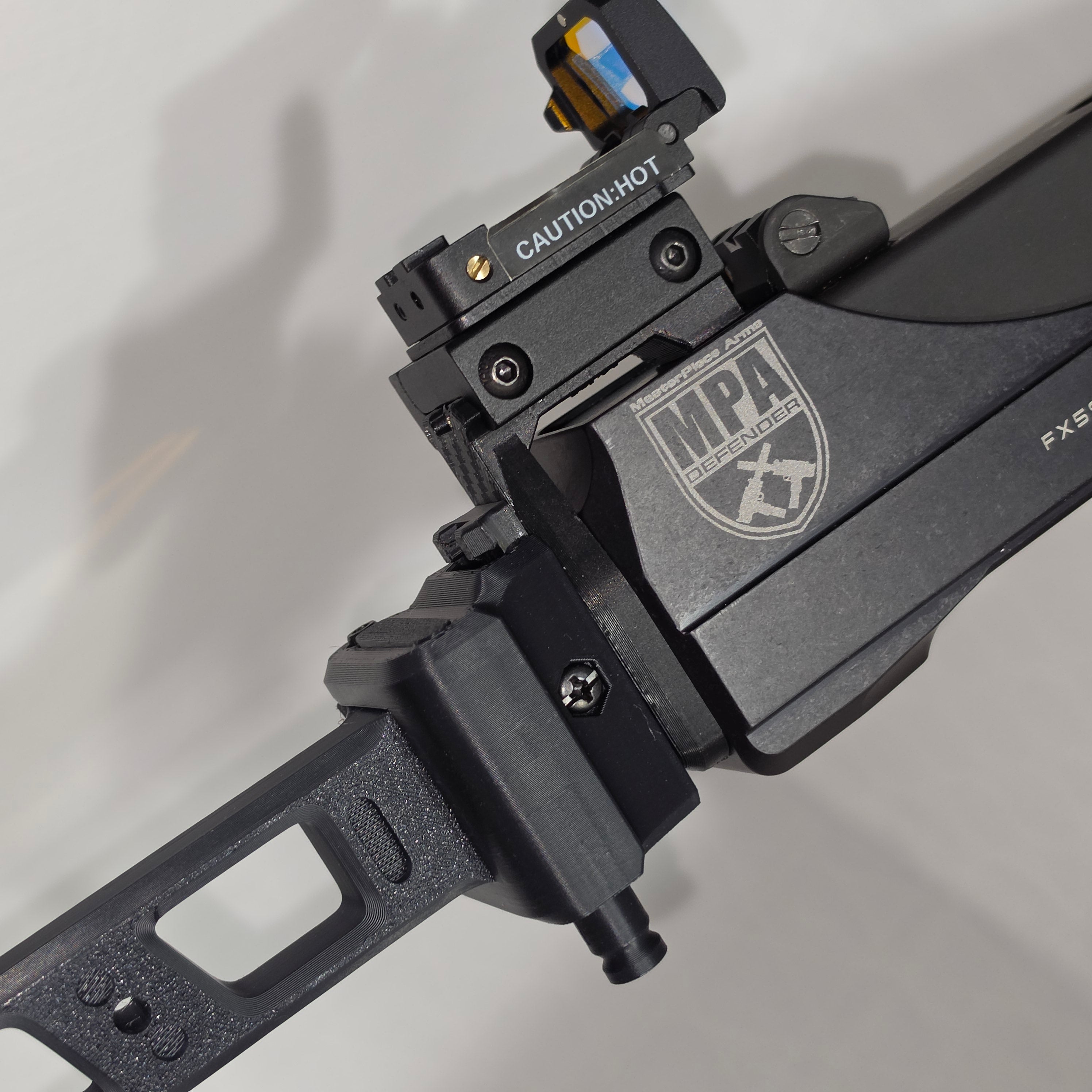 Close-up of a black tactical firearm accessory setup, MAP Defender, including a mounted red dot sight, photographed on a light neutral background.