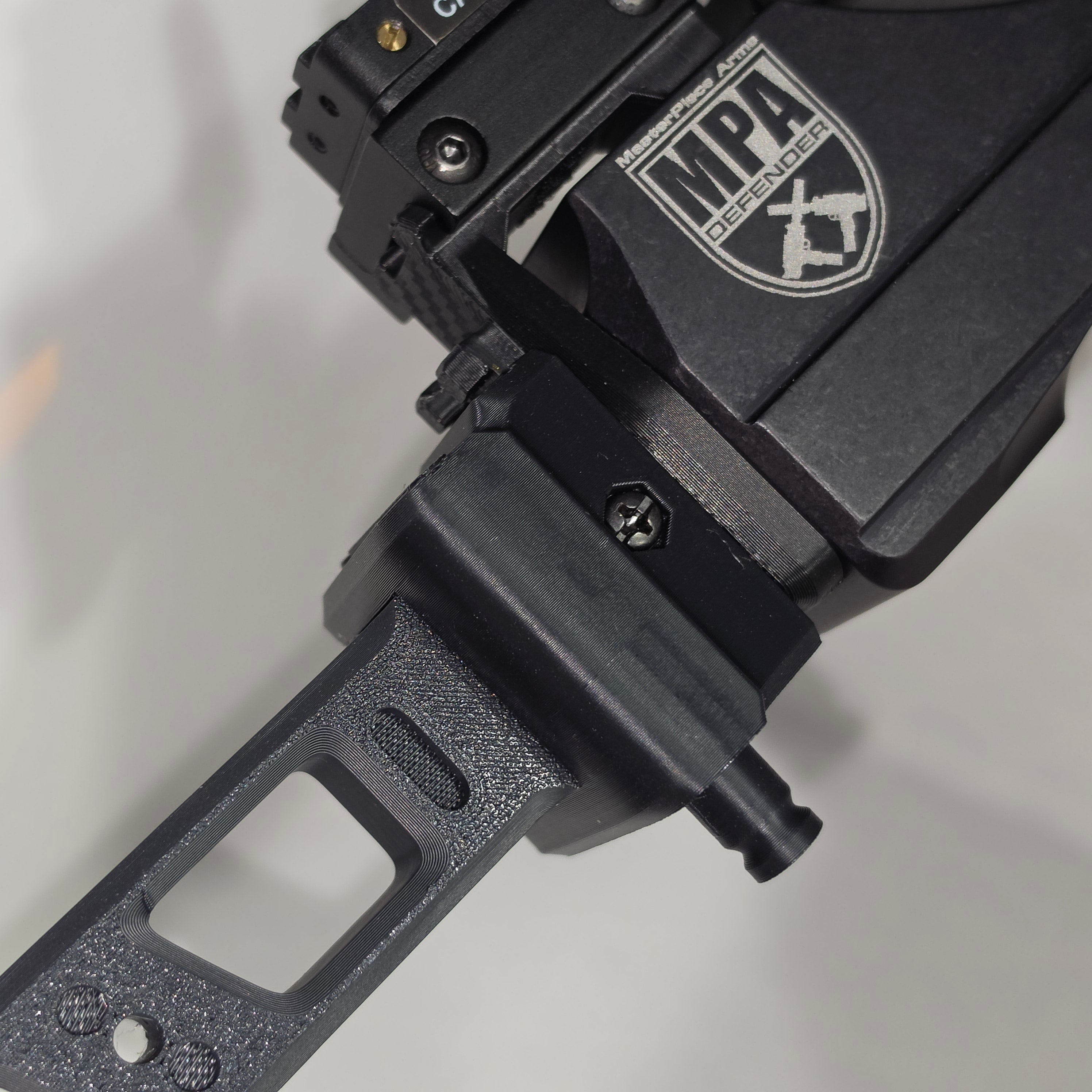 Close-up of a black tactical optic or firearm accessory with an MPA Defender, mounted on a black rail with a textured black handle or grip below.